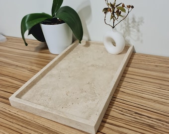 Op de afbeelding: Een rechthoekig, beige stenen dienblad met opstaande randen. Een kleine witte vaas met een cirkelvormige opening bevat gedroogde stelen. Een orchidee in pot met groene bladeren staat op de achtergrond. De lade staat op een houten oppervlak.