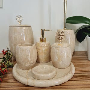 May include: A set of five beige marble bathroom accessories with gold snowflake accents. The set includes a soap dispenser, toothbrush holder, tumbler, and two jars. The accessories are arranged on a round beige marble tray.