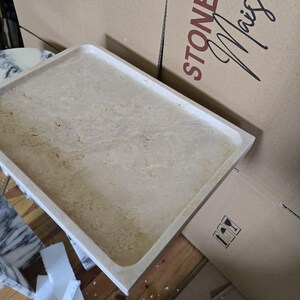 Puede incluir: Una bandeja rectangular de piedra beige con esquinas redondeadas. La bandeja tiene una superficie texturizada con vetas sutiles. Las palabras "STONE Mais" son visibles en una caja de cart&oacute;n en el fondo. La bandeja es probablemente para uso decorativo o funcional.