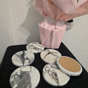 K&ouml;nnte beinhalten: Ein Set aus sechs runden Marmoruntersetzern mit einem wei&szlig;-grauen Adernmuster. Ein Untersetzer ist umgedreht und zeigt eine Korkbasis. Im Hintergrund befindet sich eine rosa Geschenkt&uuml;te.