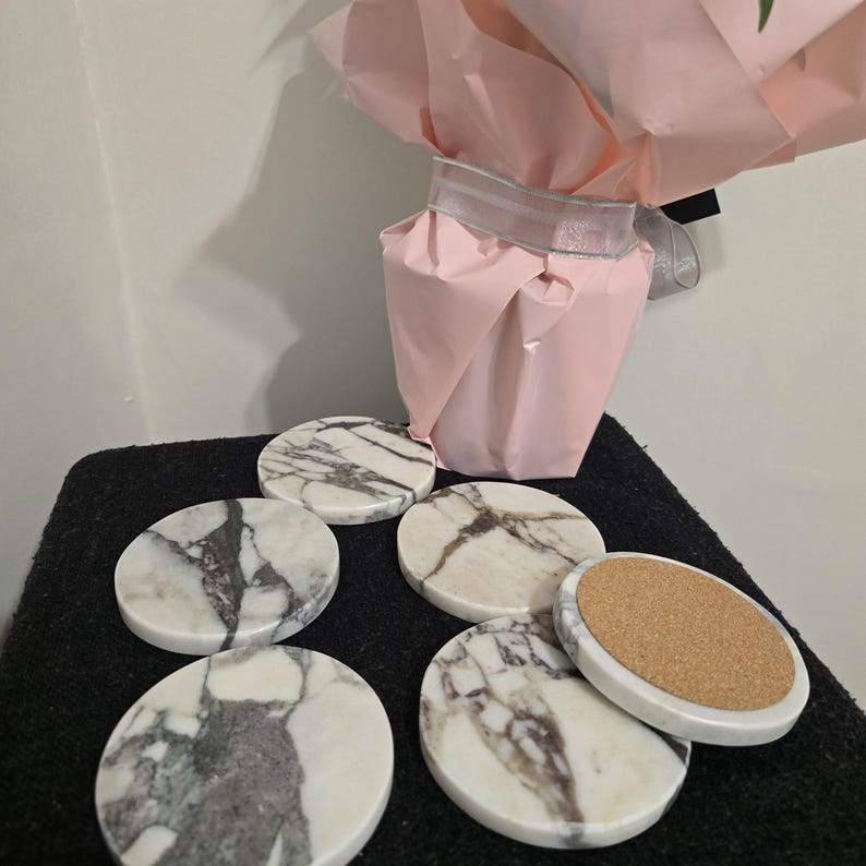 K&ouml;nnte beinhalten: Ein Set aus sechs runden Marmoruntersetzern mit einem wei&szlig;-grauen Adernmuster. Ein Untersetzer ist umgedreht und zeigt eine Korkbasis. Im Hintergrund befindet sich eine rosa Geschenkt&uuml;te.