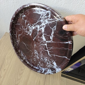 Puede incluir: Una bandeja circular con una base de color marrón rojizo intenso y vetas blancas prominentes en toda la superficie. La bandeja tiene un acabado liso y pulido con un borde exterior elevado, mostrando una apariencia similar a la piedra natural.
