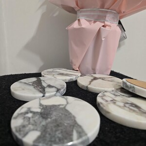 K&ouml;nnte beinhalten: Eine Sammlung runder Marmoruntersetzer mit grauer Maserung. Die Untersetzer sind auf einer dunklen Oberfl&auml;che angeordnet, im Hintergrund befindet sich ein rosa papierverpackter Gegenstand. Die Untersetzer haben einen Durchmesser von etwa 10 cm.