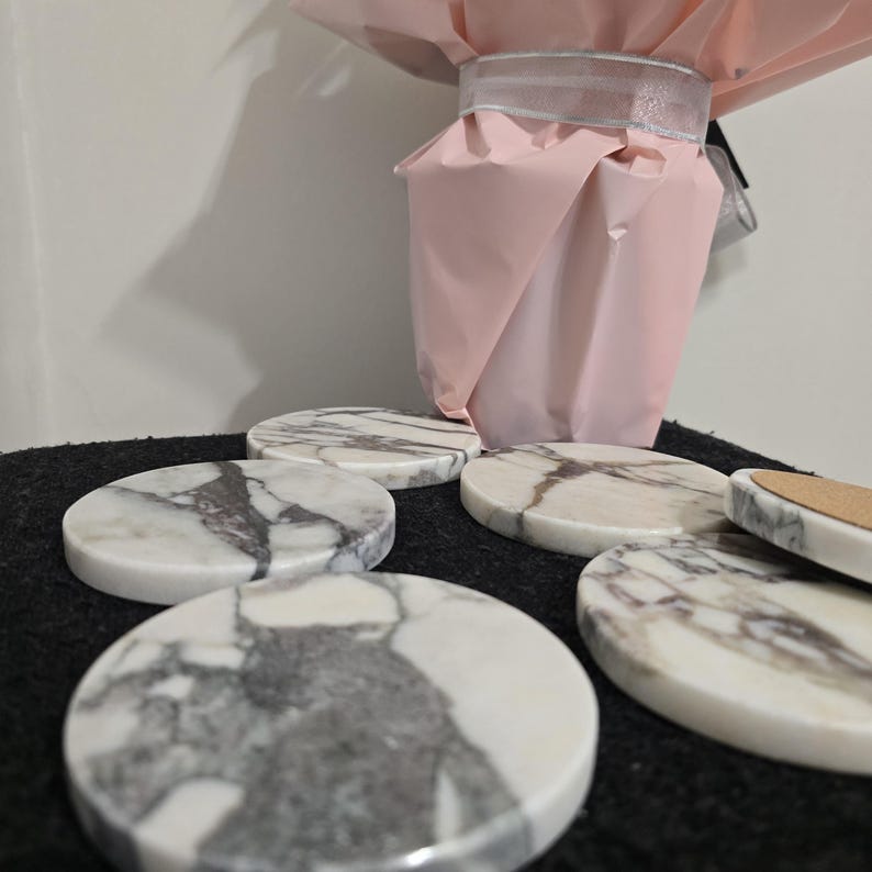 K&ouml;nnte beinhalten: Eine Sammlung runder Marmoruntersetzer mit grauer Maserung. Die Untersetzer sind auf einer dunklen Oberfl&auml;che angeordnet, im Hintergrund befindet sich ein rosa papierverpackter Gegenstand. Die Untersetzer haben einen Durchmesser von etwa 10 cm.