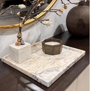 May include: A rectangular marble tray with a light brown and white veined pattern. An octagonal bronze candle holder and a white marble cube with a decorative object are on the tray. A decorative branch with white flowers is in the background.