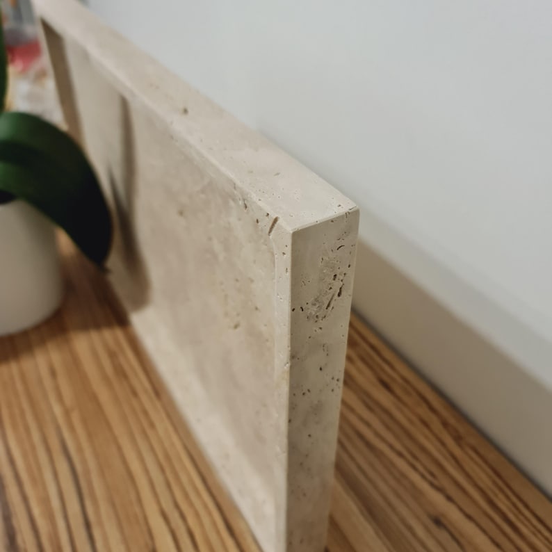 Puede incluir: Una bandeja rectangular de hormig&oacute;n beige claro con una superficie texturizada. La bandeja est&aacute; colocada contra una pared blanca y una superficie de madera. La esquina de la bandeja est&aacute; enfocada, resaltando sus l&iacute;neas limpias y su dise&ntilde;o minimalista.