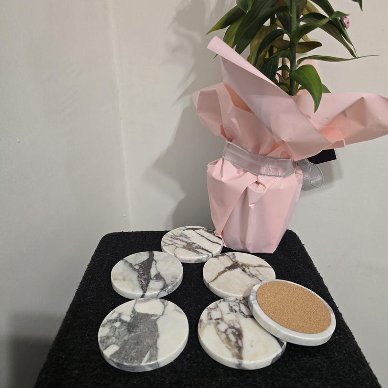 K&ouml;nnte beinhalten: Ein Set aus sechs runden Marmoruntersetzern mit grau-wei&szlig;er Maserung. Ein Untersetzer zeigt eine Korkr&uuml;ckseite. Die Untersetzer sind auf einer schwarzen Oberfl&auml;che angeordnet, mit einer rosa verpackten Pflanze im Hintergrund.