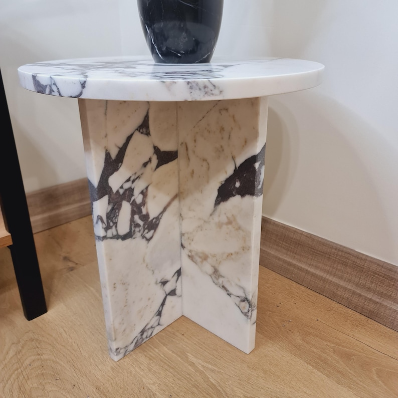 May include: A white marble side table with a round top and a unique X-shaped base. The table has a white marble top and a white marble base with gray and black veining.