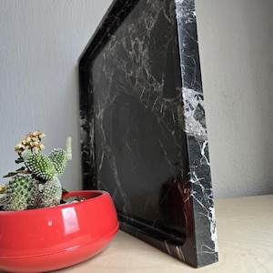 Op de afbeelding: Een vierkant zwart marmeren dienblad met opvallende witte aders, een glad gepolijst oppervlak en dikke randen. Naast het dienblad staat een kleine, felrode ronde plantenbak met een groene cactus met getextureerde stekels.