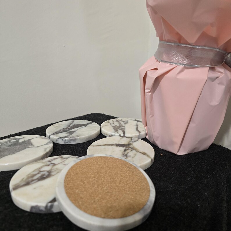 K&ouml;nnte beinhalten: Sechs runde Untersetzer aus Marmor mit grauer Maserung. Jeder Untersetzer hat eine wei&szlig;e Marmoroberfl&auml;che und einen Korkboden. Im Hintergrund befindet sich eine rosa Geschenkverpackung, die mit einem silbernen Band gebunden ist. Die Untersetzer liegen auf einer schwarzen Oberfl&auml;che.