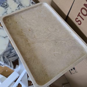 Puede incluir: Una bandeja rectangular de piedra beige con bordes redondeados. La bandeja tiene un aspecto natural veteado con sutiles variaciones de color y textura. La bandeja probablemente se utiliza para servir o exhibir.