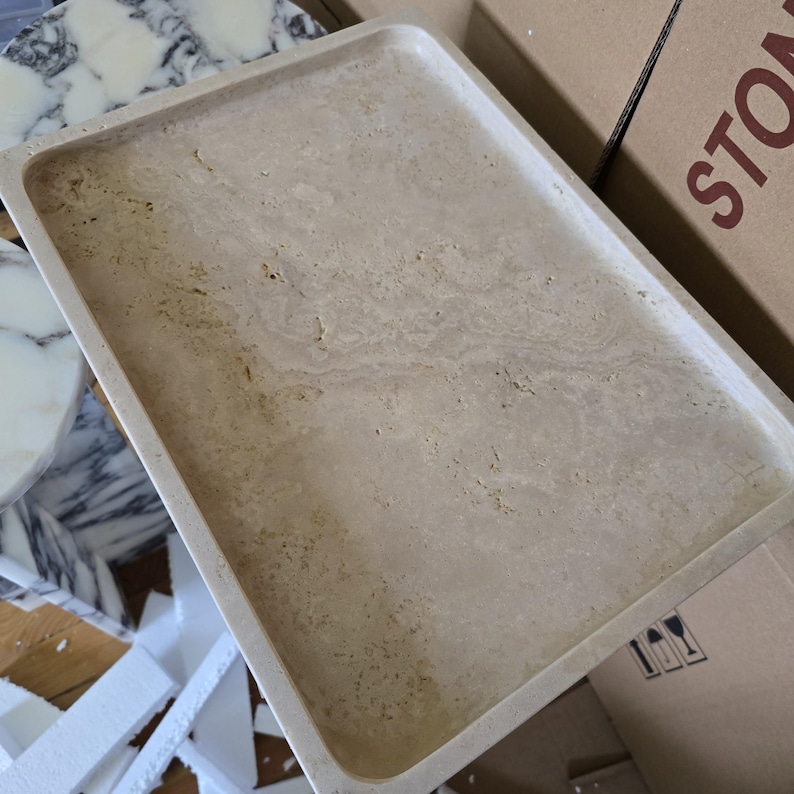 Puede incluir: Una bandeja rectangular de piedra beige con bordes redondeados. La bandeja tiene un aspecto natural veteado con sutiles variaciones de color y textura. La bandeja probablemente se utiliza para servir o exhibir.