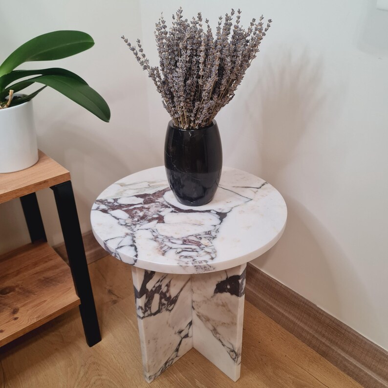 May include: A white marble side table with a round top and a unique, geometric base. The table has a black vase with dried lavender flowers on top.