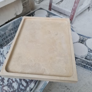 Puede incluir: Una bandeja rectangular de piedra beige con un borde elevado. La bandeja tiene una superficie lisa y probablemente est&aacute; hecha de piedra natural. La bandeja se muestra en un taller, con otras piezas de piedra visibles en el fondo.