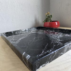 Puede incluir: Una bandeja cuadrada con una superficie negra y vetas blancas prominentes. La bandeja tiene bordes elevados y un acabado liso. El patrón oscuro similar al mármol crea un aspecto de alto contraste en la base plana y en las paredes laterales.