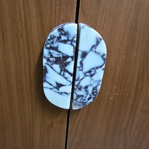 Może przedstawiać: Dwa pasujące uchwyty do szafek, każdy w kształcie półowalu, zamontowane na drewnianych drzwiach szafki. Uchwyty są w kolorze jasnoniebieskim z ciemnoszarym żyłkowaniem, przypominającym marmur. Drzwi szafki są w ciepłym, średnim brązie.