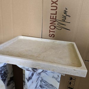 Puede incluir: Una bandeja rectangular de piedra beige con esquinas redondeadas. La bandeja se asienta sobre una base con vetas de m&aacute;rmol y blanca. El fondo presenta una caja de cart&oacute;n con el texto "STONELUX Maison".
