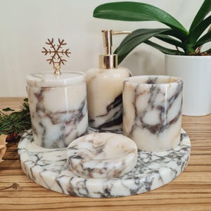 Könnte beinhalten: Ein Set aus vier Badezimmeraccessoires aus Marmor, bestehend aus einem Seifenspender, einem Glas mit einem Schneeflocken-Deckel, einem Zahnbürstenhalter und einer Seifenschale. Die Accessoires sind alle aus weißem Marmor mit grauen und braunen Adern. Das Set ist auf einem runden Marmortablett angeordnet.