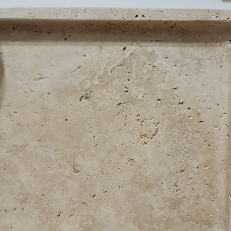 Puede incluir: Una bandeja rectangular de color beige claro con un borde ligeramente elevado. La bandeja tiene una apariencia de piedra natural con texturas visibles y peque&ntilde;os agujeros, lo que sugiere travertino o un material similar. La superficie es de tono claro y neutro.