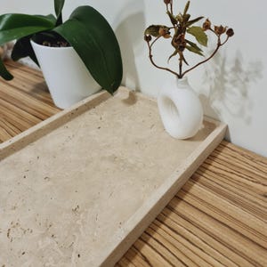 Puede incluir: Una bandeja rectangular de piedra beige con bordes elevados. Un peque&ntilde;o jarr&oacute;n blanco con un dise&ntilde;o circular &uacute;nico contiene flores secas. Tambi&eacute;n se ve una planta en maceta blanca con hojas verdes.