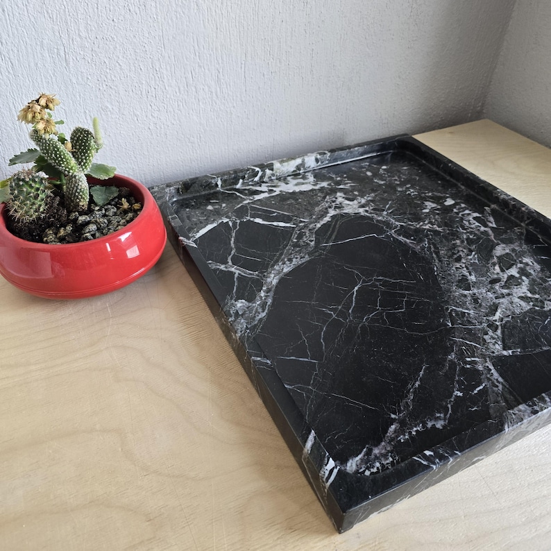 Op de afbeelding: Een vierkant zwart marmeren dienblad met opvallende witte aders en opstaande randen, naast een kleine groene cactus geplant in een gladde, glanzende rode keramische kom met een afgeronde basis en een brede opening.
