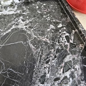 Op de afbeelding: Een rechthoekig dienblad met een donkerzwart stenen oppervlak met ingewikkelde witte en grijze aders. Het dienblad heeft een opstaande rand en een gepolijste afwerking, wat een natuurlijke marmertextuur met hoekige steenfragmenten toont.