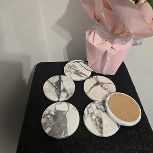 K&ouml;nnte beinhalten: Ein Set aus sechs runden Untersetzern mit wei&szlig;em Marmordesign und grauer Maserung. Ein Untersetzer zeigt eine Korkr&uuml;ckseite. Die Untersetzer sind auf einer schwarzen Oberfl&auml;che angeordnet, mit einer rosa Geschenkt&uuml;te im Hintergrund.