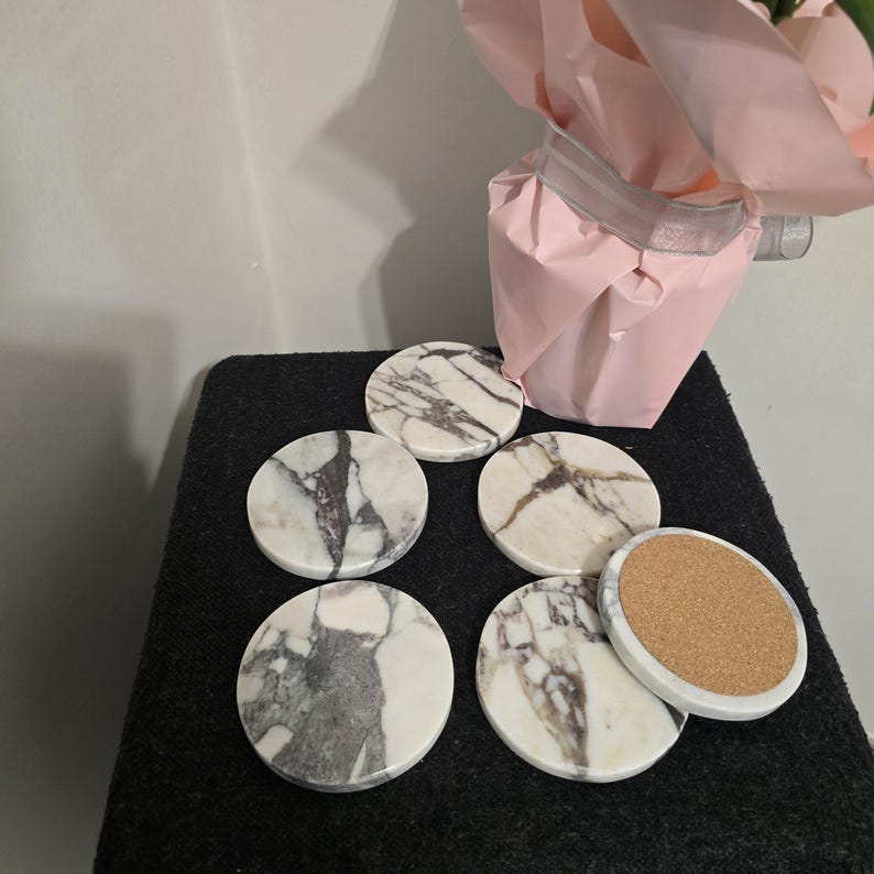 K&ouml;nnte beinhalten: Ein Set aus sechs runden Untersetzern mit wei&szlig;em Marmordesign und grauer Maserung. Ein Untersetzer zeigt eine Korkr&uuml;ckseite. Die Untersetzer sind auf einer schwarzen Oberfl&auml;che angeordnet, mit einer rosa Geschenkt&uuml;te im Hintergrund.
