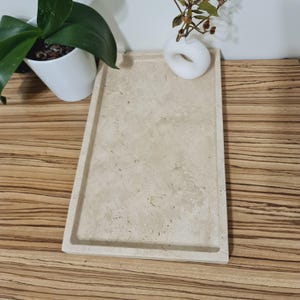 Puede incluir: Una bandeja rectangular de piedra beige con un borde ligeramente elevado. La bandeja est&aacute; sobre una superficie de madera. Un peque&ntilde;o jarr&oacute;n blanco con una planta est&aacute; en la bandeja. Una planta en maceta est&aacute; en el fondo.