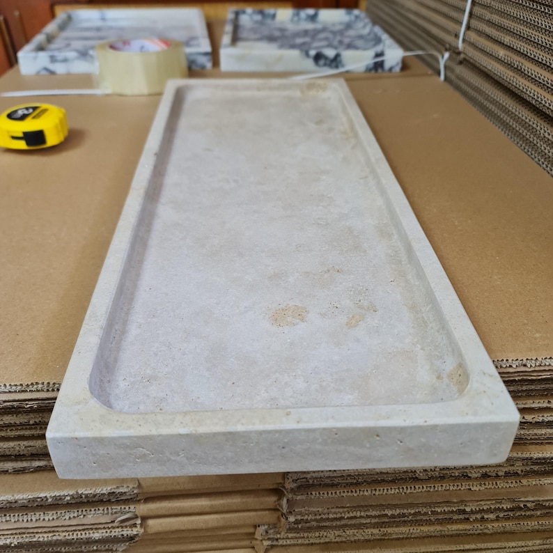 Puede incluir: Una bandeja rectangular de piedra beige claro con un borde ligeramente elevado. La bandeja tiene una superficie lisa y esquinas redondeadas. Un flex&oacute;metro amarillo es visible en el fondo, junto con otras bandejas y cart&oacute;n.
