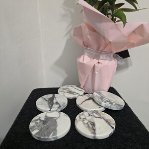 K&ouml;nnte beinhalten: Eine Sammlung von sechs runden Untersetzern aus Marmor mit grauer und wei&szlig;er Maserung. Die Untersetzer sind auf einer dunklen Oberfl&auml;che angeordnet, im Hintergrund eine Topfpflanze in rosa Papier verpackt.