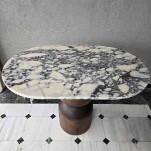 May include: Oval marble-topped table with a dark wood base. The marble surface features a white background with gray and black veining. The base is a dark, cylindrical shape. The table is a stylish piece of furniture.