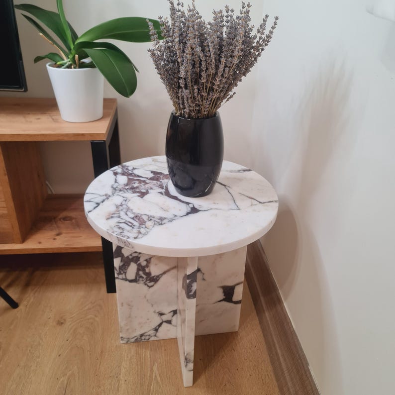 May include: A white marble side table with a round top and four legs. The table has a unique design with the legs forming a cross shape. A black vase with dried lavender flowers sits on top of the table.