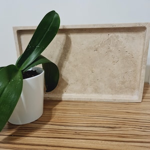 Puede incluir: Una bandeja rectangular de piedra beige con una superficie texturizada. A la izquierda de la bandeja hay una maceta de cer&aacute;mica blanca con una planta verde. La bandeja est&aacute; sobre una superficie de madera.