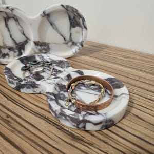 May include: A white marble jewelry tray with gray veining, holding a silver ring, a bracelet with blue accents, and a copper bracelet. Another similar tray is in the background. The trays are on a wooden surface.