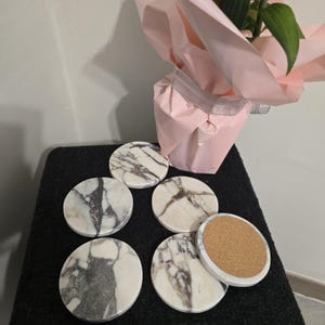 K&ouml;nnte beinhalten: Sechs runde Marmoruntersetzer mit wei&szlig;en und grauen Maserungen. Ein Untersetzer ist umgedreht und zeigt einen Korkboden. Die Untersetzer liegen auf einer schwarzen Oberfl&auml;che in der N&auml;he einer rosa verpackten Pflanze.