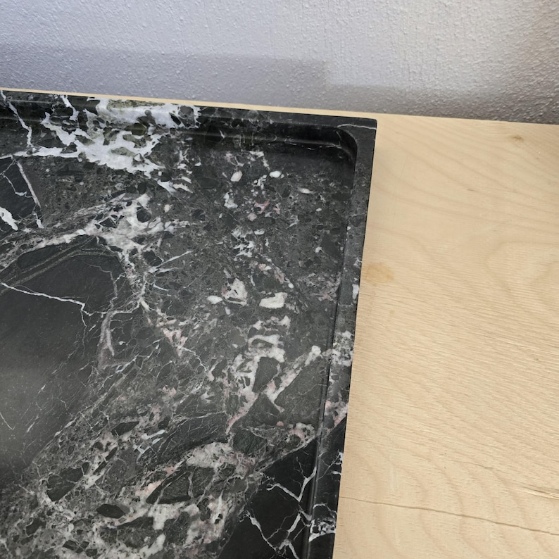 Op de afbeelding: Een rechthoekig dienblad van zwart marmer met opvallende witte aders over het gehele oppervlak. Het dienblad heeft een gladde, gepolijste afwerking met een opstaande rand die de omtrek van het donkere steenmateriaal definieert.