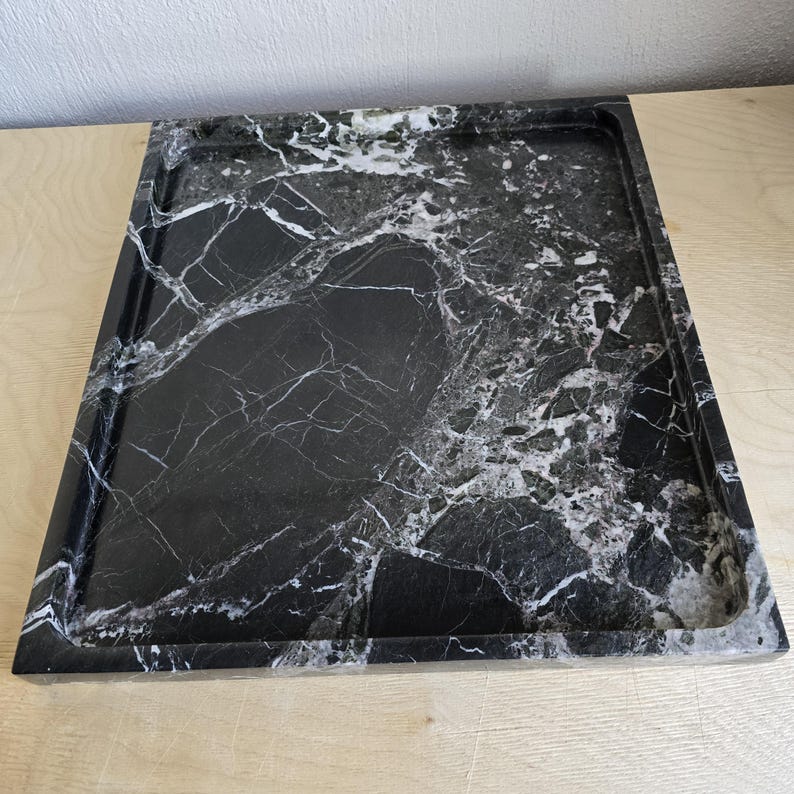 Op de afbeelding: Een rechthoekig dienblad van zwart marmer met opvallende witte aders over het gehele oppervlak. Het dienblad heeft een gladde, gepolijste afwerking met opstaande randen die een gedefinieerde omlijsting vormen en de natuurlijke steentextuur tonen.