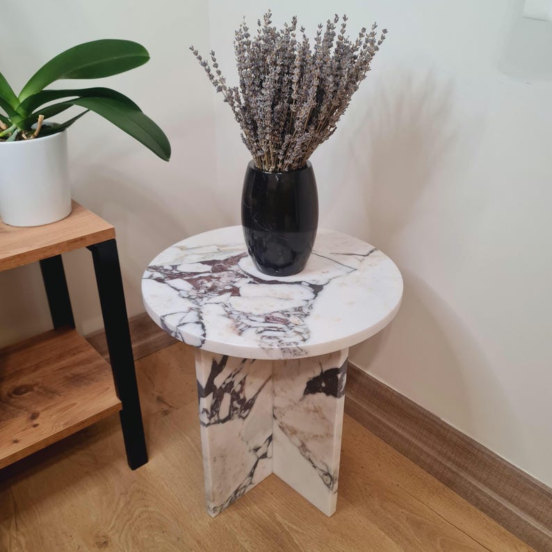 May include: A white marble side table with a round top and a cross-shaped base. The table is decorated with a natural marble pattern. A vase with dried lavender is on top of the table.