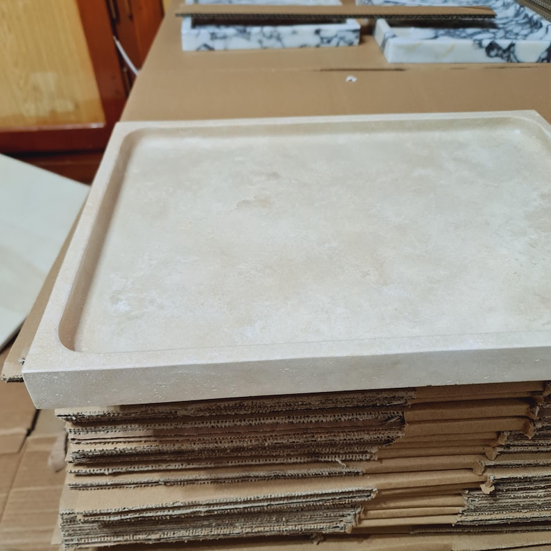 Puede incluir: Una bandeja rectangular de piedra beige con un borde ligeramente elevado. La bandeja tiene una superficie lisa y est&aacute; sobre una pila de cajas de cart&oacute;n. La piedra tiene un aspecto natural y moteado.