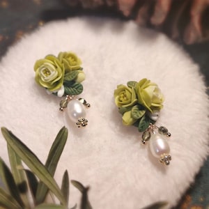 Puede incluir: Un par de pendientes florales verdes y blancos con detalles de perlas. Los pendientes presentan un grupo de tres flores con hojas verdes y una sola perla que cuelga de cada pendiente.