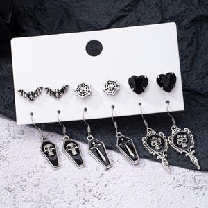 May include: A set of 8 silver tone earrings with black accents. The earrings feature a variety of designs, including bats, spiderwebs, hearts, skulls, and swords. The earrings are displayed on a white card.