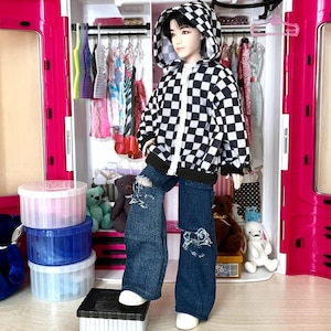 Peut inclure: Une poupée portant un sweat à capuche à carreaux noir et blanc avec une fermeture éclair, un jean bleu avec des déchirures et des baskets blanches. La poupée est debout devant un placard de maison de poupée rose et blanc.