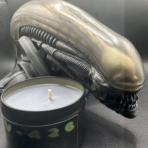 May include: A black candle in a tin with the text "V-426" and a green "6" next to it. A metallic alien head sculpture is behind the candle. The candle has a grey wax and a white wick.
