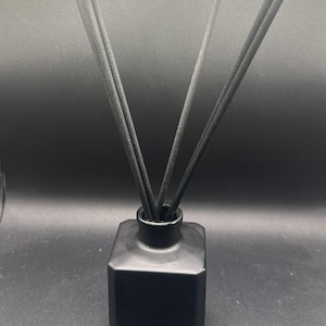 Könnte beinhalten: Ein schwarzer, quadratischer Diffusor für ätherische Öle mit mehreren schwarzen Schilfrohren. Der Diffusor hat einen schmalen Hals und eine breite Basis. Die Schilfrohre sind fächerförmig angeordnet und erstrecken sich nach oben aus der Flasche.
