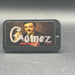 Op de afbeelding: Een zwarte rechthoekige doos met een vintage portret van een man met een snor, die een sigaar vasthoudt. De naam "Gomez" staat in een gestileerd lettertype. De tekst "Phantom Foundry Co." en "0,5 oz / 14,17 g" zijn ook zichtbaar.