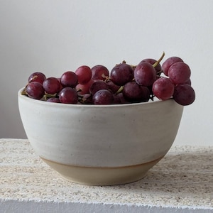 May include: A white ceramic bowl with a light brown rim, filled with red grapes.