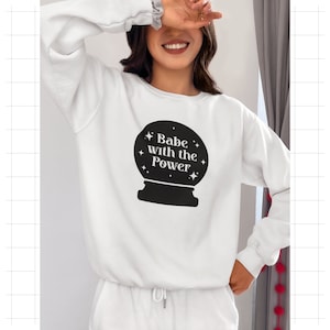 Può includere: Felpa bianca con la scritta "Babe with the Power" in una grafica nera a sfera di cristallo. La felpa fa parte di un completo coordinato. Le maniche hanno un polsino arricciato.
