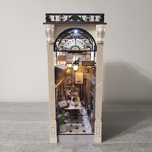 May include: A miniature diorama book nook featuring a Parisian-style street scene with a black arched metal frame, white pillars, and a black railing. The interior displays a chequered floor, a lit lantern, tables with pink flowers, and signs reading Paris and K Book.