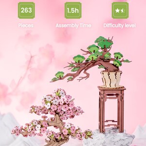 May include: Two wooden 3D puzzles of cherry blossom bonsai trees. The larger tree has green leaves and is on a stand. The smaller tree has pink blossoms and is in a pot. Both trees are on a white surface with pink flower petals.